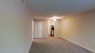This single-floor residential unit offers a straightforward and functional layout designed for comfortable everyday living. The main living area is finished with wall-to-wall carpeting in a neutral tone, creating a warm and cohesive atmosphere throughout the space. Soft, neutral-colored walls contribute to a clean and understated aesthetic that allows for flexible interior styling.

A ceiling fan is centrally positioned in the living area, providing both overhead lighting and air circulation. The open sightlines from the living area lead directly into the kitchen, which is compact yet well-equipped with stainless steel appliances, including a refrigerator and range. The kitchen features hard flooring, offering a practical contrast to the carpeted living space.

A white interior door is visible along one wall, providing access to an additional room or closet, contributing to the unit's overall utility. Floor-level vents indicate a forced-air climate control system throughout. The layout is efficient and uncluttered, making it easy to navigate and personalize. Overall, the space presents a tidy, move-in-ready environment suited to a range of lifestyles, with all essential features thoughtfully integrated into a compact, single-level footprint.