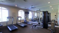 This single-story fitness space is designed to support a well-rounded exercise routine within a compact, functional layout. The room features wall-to-wall mirrors along one side, which visually expand the space and allow users to monitor their form during workouts. A ceiling fan is centrally mounted to provide air circulation during exercise sessions.

Natural light enters through multiple arched windows, lending the room a bright and open feel while offering views of the surrounding outdoor greenery. The windows are fitted with blinds for adjustable light control. Recessed lighting supplements the natural light, ensuring the space remains well-lit at all hours.

The fitness equipment includes a treadmill and an elliptical trainer for cardiovascular workouts, along with a multi-function cable and weight machine for strength training. A flat-screen television is mounted on the wall, providing entertainment during cardio sessions. A workout bench is also present, adding versatility for strength exercises.

The flooring is covered in durable, dark carpeting suited for a fitness environment. Crown molding along the ceiling adds a subtle architectural detail, giving the room a polished finish. An informational bulletin board is mounted near the entrance for user reference.