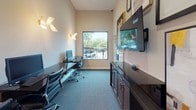 This single-floor business center is thoughtfully designed to provide a functional and visually engaging workspace. The room features patterned blue-grey carpeting that adds texture underfoot, while neutral walls provide a clean backdrop throughout. A large picture window floods the space with natural light and offers views of the surrounding outdoor area, including mature trees and a parking area beyond.

Multiple sleek, dark workstations line the walls, each equipped with desktop computers, keyboards, and ergonomic black leather task chairs, accommodating several users simultaneously. Warm-toned wall sconces with gold brass fixtures supplement natural light, adding a refined touch to the otherwise practical environment. A wall-mounted flat-screen display is positioned prominently for easy viewing, accompanied by a dark wood credenza with cabinet storage below.

The walls serve as a curated gallery, showcasing an eclectic collection of framed abstract and contemporary artworks in a coordinated palette of black, white, yellow, and gold. A glass-paneled black door connects the business center to an adjacent space, maintaining visual connectivity while defining the room's boundaries. The overall ambiance balances professional functionality with tasteful, modern design details.