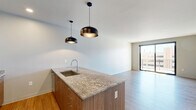 Located at 200 N Washington St in downtown Green Bay, Wisconsin, this modern one-bedroom, one-bathroom residence offers a well-designed layout across a single floor. The open-concept kitchen and living area serves as the heart of the unit, showcasing warm wood-look flooring, crisp white cabinetry, and a substantial granite-topped island with an undermount sink and stylish pendant lighting. Stainless steel appliances, including a refrigerator, range, and microwave, complete the contemporary kitchen. The living area features large sliding glass doors that lead to a private balcony, offering elevated views of a community pool and the surrounding downtown Green Bay cityscape. The bedroom is carpeted for comfort, features a wall-mounted climate control unit, and includes a large window that brings in generous natural light with additional urban views. Built-in closet storage flanks the entrance to the bathroom, which is finished with tile flooring, a wood-toned vanity with granite countertop, and a walk-in glass-enclosed shower. A dedicated laundry closet with stacked washer and dryer units adds everyday convenience. The entryway features a bold dark front door alongside a full-height built-in storage unit, establishing a clean and functional arrival experience throughout.