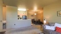This single-floor space offers an open-concept layout that thoughtfully integrates the main living areas into one cohesive environment. The kitchen is positioned behind a half-wall breakfast bar, providing a natural division between the cooking and living spaces while maintaining an open, airy feel. It features light wood cabinetry, a black refrigerator, and ample counter space, making it both functional and accessible. A coffee station adds a practical, homey touch to the kitchen setup.

Adjacent to the kitchen, the dining area comfortably accommodates a dark wood table and chair set, perfectly suited for everyday meals or entertaining guests. A pendant light fixture hangs overhead, providing focused illumination and adding a decorative element to the space. A charming "Bon Appétit" wall sign accents the dining nook, reinforcing a warm, welcoming atmosphere.

The living area flows naturally from the dining space, featuring plush carpeted flooring that adds comfort underfoot. Neutral, warm-toned walls throughout create a cohesive and inviting ambiance. The textured ceilings and recessed lighting contribute to the overall warmth of the interior. Together, these spaces form a practical and comfortable single-floor layout ideal for everyday living.