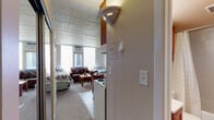 This single-floor unit offers a practical and efficient layout designed to maximize every square foot. The main living area serves as the heart of the space, combining a sleeping zone and a lounge area in one open room. Large windows fitted with vertical blinds allow natural light to fill the carpeted space, while a wall-mounted air conditioning unit provides climate control. A cozy sofa, coffee table, and bed are comfortably arranged within this shared room, creating a versatile and functional living environment.

Adjacent to the living area is the kitchen, which is well-equipped with warm wood cabinetry extending to the ceiling for generous storage. It features a full-size refrigerator, an electric coil range with overhead ventilation, a sink, and countertop appliances including a microwave. A ceiling fan with a light fixture keeps the space comfortable. A round dining table with seating adds a dedicated area for meals.

The bathroom is clean and straightforward, outfitted with a single-basin vanity, a toilet, and a tub-shower combination enclosed by a curtain. Wood-toned cabinetry above provides additional storage. Tile flooring completes the practical design. Throughout the unit, a consistent use of neutral tones and warm wood finishes creates a cohesive, understated aesthetic suited for comfortable day-to-day living.