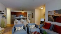 This single-floor residence showcases an open-concept design that seamlessly connects the kitchen and living areas, creating a cohesive and functional layout. The kitchen is outfitted with rich, warm-toned wood cabinetry, stainless steel appliances including a refrigerator, range, and microwave, and a dark-surfaced breakfast bar with modern white bar stools. Track lighting illuminates the kitchen workspace, highlighting the contemporary finishes throughout.

The living area flows naturally from the kitchen, furnished with plush, blue-gray upholstered seating accented by decorative patterned throw pillows in muted tones. A floor lamp provides ambient lighting, complementing the warm overhead fixtures. Neutral-toned walls and light flooring create a bright, airy atmosphere throughout the space. A partial view into an adjacent hallway or secondary room suggests additional functional areas beyond the main living zone. Decorative wall accents and potted greenery add personality and warmth. Overall, the space presents a balanced combination of practicality and modern comfort, well-suited for everyday living and casual entertaining.