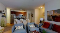 This single-floor residence showcases an open-concept design that seamlessly connects the kitchen and living areas, creating a cohesive and functional layout. The kitchen is outfitted with rich, warm-toned wood cabinetry, stainless steel appliances including a refrigerator, range, and microwave, and a dark-surfaced breakfast bar with modern white bar stools. Track lighting illuminates the kitchen workspace, highlighting the contemporary finishes throughout.

The living area flows naturally from the kitchen, furnished with plush, blue-gray upholstered seating accented by decorative patterned throw pillows in muted tones. A floor lamp provides ambient lighting, complementing the warm overhead fixtures. Neutral-toned walls and light flooring create a bright, airy atmosphere throughout the space. A partial view into an adjacent hallway or secondary room suggests additional functional areas beyond the main living zone. Decorative wall accents and potted greenery add personality and warmth. Overall, the space presents a balanced combination of practicality and modern comfort, well-suited for everyday living and casual entertaining.