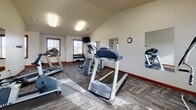 This fitness center is thoughtfully designed to support a full range of workout routines within a single, open floor plan. The space features vaulted ceilings that lend an open, airy feel, while large windows with slatted blinds allow natural light to stream across the textured, multi-tone carpet flooring throughout. Warm red-brown baseboards and trim provide a consistent, polished finish that contrasts nicely with the neutral walls.

The main exercise area is well-appointed with multiple treadmills, an elliptical trainer, a stationary bike, and a cable machine, offering a comprehensive selection of cardio and strength equipment. A dedicated free-weight station includes a full dumbbell rack, accompanied by an adjustable workout bench and floor mat for strength training and stretching. Wall-mounted mirrors are strategically placed throughout to maximize the sense of space and allow users to monitor their form during exercise.

A wall-mounted flat-screen television is positioned for easy viewing during cardio workouts. A corridor connects to an adjacent room, which offers additional open floor space ideal for stretching, yoga, or floor exercises — further complemented by mirrors and a mounted television. A coat hook rail near the hallway entrance adds a practical touch. The overall ambiance is clean, functional, and well-lit, making this fitness center a practical and inviting space for daily use.