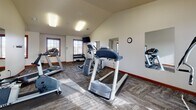 This fitness center is thoughtfully designed to support a full range of workout routines within a single, open floor plan. The space features vaulted ceilings that lend an open, airy feel, while large windows with slatted blinds allow natural light to stream across the textured, multi-tone carpet flooring throughout. Warm red-brown baseboards and trim provide a consistent, polished finish that contrasts nicely with the neutral walls.

The main exercise area is well-appointed with multiple treadmills, an elliptical trainer, a stationary bike, and a cable machine, offering a comprehensive selection of cardio and strength equipment. A dedicated free-weight station includes a full dumbbell rack, accompanied by an adjustable workout bench and floor mat for strength training and stretching. Wall-mounted mirrors are strategically placed throughout to maximize the sense of space and allow users to monitor their form during exercise.

A wall-mounted flat-screen television is positioned for easy viewing during cardio workouts. A corridor connects to an adjacent room, which offers additional open floor space ideal for stretching, yoga, or floor exercises — further complemented by mirrors and a mounted television. A coat hook rail near the hallway entrance adds a practical touch. The overall ambiance is clean, functional, and well-lit, making this fitness center a practical and inviting space for daily use.