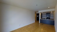 This single-floor space showcases an open-concept layout that seamlessly connects the main living area to the kitchen. The living area is characterized by smooth white walls and warm hardwood flooring that extends throughout, adding a natural, understated tone to the interior. The clean white ceiling with recessed lighting provides even illumination across the space.

Transitioning toward the back, a kitchen area becomes visible, distinguished by a dark-toned counter or bar surface that provides a subtle contrast against the lighter surroundings. Open wall-mounted shelving units flank the kitchen zone, offering practical storage and display space. A black refrigerator anchors the kitchen area, and recessed lighting continues overhead, maintaining a consistent lighting scheme.

The overall design aesthetic is minimal and functional, with neutral tones dominating the palette. The exposed wood flooring throughout gives the space a warm, tactile quality, while the open layout encourages flexible use and arrangement. The straightforward architectural detailing and absence of ornamental features lend the space a clean, contemporary character well-suited to a variety of uses.