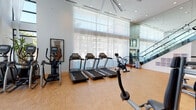 Located at 22121 Clarendon St in Woodland Hills, Los Angeles, this well-equipped fitness center offers a bright, modern environment designed to support a comprehensive workout experience. The facility features soaring double-height ceilings and expansive floor-to-ceiling windows dressed with sheer curtains, flooding the space with abundant natural light and creating an open, airy atmosphere.

The main workout floor is finished with durable cork flooring throughout and is organized into distinct exercise zones. The cardio area is equipped with multiple treadmills, elliptical trainers, and stationary bikes, all positioned to take advantage of the natural light from the large windows. The strength training section includes a variety of selectorized weight machines and cable systems, as well as a free weights area stocked with a full rack of dumbbells. A dedicated functional fitness zone houses stability balls and accessories for flexibility and core training.

A striking glass-railed staircase serves as an architectural focal point, leading to an upper-level mezzanine observation area, reinforcing the facility's modern, upscale design aesthetic. Floor-to-ceiling mirrors are strategically placed throughout to enhance the sense of space and support proper form during exercise. Recessed lighting complements the natural light, ensuring the space remains well-lit at all times. The overall ambiance is clean, contemporary, and professional.