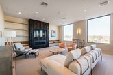 This contemporary amenity space is thoughtfully designed to serve as a vibrant communal hub, offering a variety of distinct zones that cater to socializing, relaxing, and gathering. The open layout flows seamlessly between areas, each with its own character while maintaining a cohesive modern aesthetic.

The main lounge area features plush, neutral-toned sofas and rounded accent chairs in warm terracotta hues, centered around a striking dark fireplace surround with integrated built-in shelving and a mounted television. Recessed lighting and sleek wall sconces provide warm, layered illumination throughout.

A full-service bar and kitchen area showcases bold design choices, including a dramatic geometric-patterned accent wall in coral, charcoal, and cream tones, dark granite countertops, and dark cabinetry. A built-in draft beer tap system and pass-through window connect to the lounge, making it ideal for hosting events.

Tall, narrow windows line the perimeter of the space, flooding the interior with natural light and offering sweeping elevated views of the surrounding cityscape and treetops. A secondary lounge zone features herringbone-patterned light wood flooring, curated artwork, and additional seating for quieter use.

High-top bar tables positioned along the window line invite casual conversation while taking in the views. The overall ambiance is sophisticated, energetic, and welcoming.