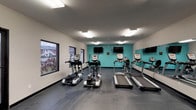 This well-equipped fitness center on Shawnee Trail offers a comprehensive workout environment designed to accommodate a wide range of exercise routines. The single-floor facility is divided into distinct functional zones, creating an organized and efficient layout for users.

The cardio area is outfitted with multiple treadmills and elliptical machines, each paired with individual mounted flat-screen televisions for entertainment during workouts. A full wall of mirrors runs along one side, enhancing the sense of space and allowing users to monitor their form. Large windows along the perimeter flood the space with natural light and provide views of the surrounding exterior.

The strength training section features a robust selection of cable machines and selectorized weight equipment, along with adjustable workout benches suitable for a variety of exercises. This area also benefits from mirror coverage, contributing to a cohesive and professional gym atmosphere.

The facility is finished with durable rubber flooring throughout, providing safety and comfort underfoot. Vibrant teal accent walls add a bold, energizing aesthetic against the neutral white surroundings. Bright overhead fluorescent lighting ensures excellent visibility across the entire space.

A dedicated utility area includes restroom access, a water fountain, and standard safety equipment, ensuring the space is both functional and compliant. Overall, the fitness center is a well-appointed and practical facility.
