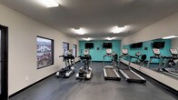 This well-equipped fitness center on Shawnee Trail offers a comprehensive workout environment designed to accommodate a wide range of exercise routines. The single-floor facility is divided into distinct functional zones, creating an organized and efficient layout for users.

The cardio area is outfitted with multiple treadmills and elliptical machines, each paired with individual mounted flat-screen televisions for entertainment during workouts. A full wall of mirrors runs along one side, enhancing the sense of space and allowing users to monitor their form. Large windows along the perimeter flood the space with natural light and provide views of the surrounding exterior.

The strength training section features a robust selection of cable machines and selectorized weight equipment, along with adjustable workout benches suitable for a variety of exercises. This area also benefits from mirror coverage, contributing to a cohesive and professional gym atmosphere.

The facility is finished with durable rubber flooring throughout, providing safety and comfort underfoot. Vibrant teal accent walls add a bold, energizing aesthetic against the neutral white surroundings. Bright overhead fluorescent lighting ensures excellent visibility across the entire space.

A dedicated utility area includes restroom access, a water fountain, and standard safety equipment, ensuring the space is both functional and compliant. Overall, the fitness center is a well-appointed and practical facility.
