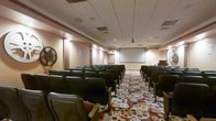 This single-floor screening room offers a purpose-built environment designed for presentations, film screenings, and group gatherings. The space is arranged in a classic theater layout, with rows of dark upholstered, fixed auditorium-style seats oriented toward a large projection screen mounted at the front of the room. A ceiling-mounted projector ensures a clear, centered image for all attendees. The drop ceiling features a grid-tile design with recessed and ambient lighting that creates a warm, inviting atmosphere throughout the room. Decorative columns line the walls, adding a touch of architectural character to the interior. The standout design element is the boldly patterned carpet, which showcases a film reel motif in earthy tones of red, brown, and cream — a clear nod to the room's cinematic purpose. A film reel wall decoration further reinforces the movie theater theme. A podium or lectern is positioned near the front, making the space equally suited for lectures and formal presentations. The overall ambiance is polished and functional, combining classic auditorium design with thematic décor to create an engaging and comfortable environment for audiences of all kinds.