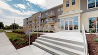 The Encore is a multi-story residential building presenting a contemporary architectural design that blends neutral-toned brick cladding with horizontal lap siding in warm gray tones. The building's façade is punctuated by private balconies equipped with metal railings, offering residents outdoor space with open views of the surrounding area. Large floor-to-ceiling windows at the main entrance flood the lobby level with natural light, creating an inviting and transparent street presence. The primary entrance is accessed via broad concrete steps complemented by a metal handrail, ensuring accessibility alongside a designated ramp approach. Thoughtfully landscaped grounds frame the building's base, featuring ornamental plantings, decorative mulched beds, and low-maintenance shrubs that add greenery and curb appeal. The exterior palette combines earthy brick tones with contemporary siding, giving the building a modern yet grounded aesthetic. Prominent signage identifies the property at the entrance corner, reinforcing its distinct identity. The overall design balances clean architectural lines with practical amenities, reflecting a well-considered approach to modern residential living in an urban setting.