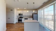 Located in Lombard, IL, this single-floor space offers a well-designed, contemporary layout that emphasizes both functionality and modern aesthetics. The open-concept kitchen serves as the centerpiece of the space, outfitted with crisp white shaker-style cabinetry extending to the ceiling for generous storage capacity. A full suite of stainless steel appliances — including a gas range, over-the-range microwave, dishwasher, and refrigerator — provides a complete, move-in-ready cooking environment. Light granite countertops run throughout the kitchen, continuing onto a central island that doubles as a breakfast bar and prep area. Dual pendant lights with warm brass-toned fixtures hang above the island, complementing the recessed lighting overhead. A tile backsplash adds a clean, finished detail to the cooking area, while vertical blind-covered sliding glass doors bring in natural light and offer outdoor access. Wood-look plank flooring runs consistently throughout the space, adding warmth and visual continuity. The bathroom features a tub-shower combination with tiled surround, granite-topped vanity, and brushed bronze hardware accents. The open living area adjacent to the kitchen provides a versatile, light-filled space well-suited for a variety of layouts and uses.