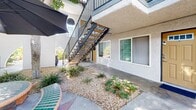 Located at 10853 Firestone Blvd in Norwalk, California, this multi-unit residential property presents a classic two-story stucco building with a clean, well-maintained exterior. The building's design features a central courtyard corridor that runs the length of the property, providing a welcoming pedestrian pathway connecting all units and common areas. Ground-floor units benefit from direct exterior entry, while upper-floor units are accessed via metal-railed staircases with wooden treads that are positioned at key points along the courtyard. Upper-level units feature private balconies enclosed by dark metal railings, offering elevated outdoor space and views over the courtyard. The courtyard landscaping incorporates drought-tolerant plantings, including agave, ornamental shrubs, and mature shade trees that line the walkway, creating a pleasant and low-maintenance outdoor environment. Decorative elements such as a large ceramic planter and stamped concrete detailing add visual interest to the shared walkway. The exterior walls feature a textured stucco finish in a light neutral tone accented by dark trim, with individual unit doors finished in warm gold tones. Windows throughout provide natural light to each unit. The overall atmosphere is calm and well-organized, offering a comfortable residential setting in a convenient Norwalk location.