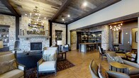 This single-story space showcases a masterful blend of rustic and refined design elements throughout its open floor plan. The reclaimed wood plank ceiling, accented by dark exposed beams, creates a warm, cabin-like atmosphere overhead, while an elegant wrought-iron chandelier adds a touch of sophistication. Underfoot, wide-plank hardwood floors in a honey tone complement the earthy palette of the space.

Anchoring the living area is a striking stone fireplace with a substantial wood mantel, flanked by a built-in niche that offers both display and storage potential. The surrounding walls feature natural stacked stone, lending authenticity and texture that ties the interior to its rustic aesthetic.

Through a stone archway supported by dark wood beams, the space opens into a well-appointed bar or kitchen area. Pendant lighting illuminates a reclaimed wood counter with bar seating, while open shelving along the back wall provides organized display space. The seamless transition between the lounge and this functional area makes the layout ideal for entertaining, offering a cohesive and inviting environment throughout.