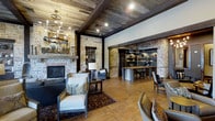 This single-story space showcases a masterful blend of rustic and refined design elements throughout its open floor plan. The reclaimed wood plank ceiling, accented by dark exposed beams, creates a warm, cabin-like atmosphere overhead, while an elegant wrought-iron chandelier adds a touch of sophistication. Underfoot, wide-plank hardwood floors in a honey tone complement the earthy palette of the space.

Anchoring the living area is a striking stone fireplace with a substantial wood mantel, flanked by a built-in niche that offers both display and storage potential. The surrounding walls feature natural stacked stone, lending authenticity and texture that ties the interior to its rustic aesthetic.

Through a stone archway supported by dark wood beams, the space opens into a well-appointed bar or kitchen area. Pendant lighting illuminates a reclaimed wood counter with bar seating, while open shelving along the back wall provides organized display space. The seamless transition between the lounge and this functional area makes the layout ideal for entertaining, offering a cohesive and inviting environment throughout.
