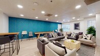 This single-floor communal space is designed to encourage socializing and relaxation through a well-balanced mix of vibrant design and functional furnishings. The main lounge area immediately draws attention with a striking teal accent wall adorned with a geometric gold-line pattern, serving as a bold focal point that energizes the room. Light wood-look flooring runs throughout, complementing the neutral tones of the gray upholstered sofas, armchairs, and accent chairs arranged around a natural wood coffee table. A wall-mounted flat-screen television is positioned within the seating area, making it suitable for group viewing. Multiple windows along the perimeter allow natural light to filter in, contributing to an open and welcoming atmosphere. A shuffleboard table is positioned along the teal accent wall, adding a recreational element to the space, while a tall bar-height table with stools offers a casual seating alternative. An adjacent kitchenette provides practical amenities, featuring white subway tile backsplash, open shelving, white cabinetry, a sink, and a compact countertop appliance area paired with a butcher-block high table and seating. Recessed lighting throughout ensures consistent illumination. The overall ambiance is modern and inviting, balancing playful design elements with comfortable, communal-focused functionality.