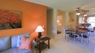 This single-floor residence offers a well-connected open layout designed for comfortable everyday living. The living area immediately draws attention with a bold accent wall in a warm burnt orange hue, creating a striking focal point that adds character and energy to the space. Light-toned carpeting runs throughout, contributing to a clean and inviting atmosphere. The living area is thoughtfully arranged to flow seamlessly into an adjacent dining space, where a dark metal-framed dining set provides a stylish contrast to the softer surroundings. Beyond the dining area, the kitchen is visible through an open passageway, featuring white cabinetry, light countertops, and tile flooring — offering a bright, functional workspace. The kitchen's white finishes reflect light effectively, keeping the area feeling open and airy. The overall color palette balances warm and neutral tones, from the orange accent wall to the beige and cream finishes found throughout the common areas. The single-floor layout ensures easy accessibility and a cohesive sense of continuity from room to room. This space combines practical design with personality-driven details, making it both functional and visually engaging for daily life.