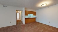 This single-floor, two-bedroom, one-bathroom unit offers a practical and straightforward layout well-suited for everyday living. The living area is carpeted in warm beige tones and features a textured ceiling, with a sliding glass door that opens to a private balcony overlooking lush greenery, bringing in natural light and outdoor views. A half-wall pass-through connects the living space to the kitchen, which is designed in a galley configuration. The kitchen features white cabinetry with wood-tone trim, white appliances including a refrigerator, electric range, and dishwasher, as well as a double sink and dark hardwood-style flooring that adds contrast to the bright cabinetry. Both bedrooms are generously sized, carpeted, and include windows with blinds that allow natural light. One bedroom also features a closet with built-in shelving for added storage. The bathroom is finished with wood-style flooring, a wide vanity with white countertop and a large mirror, and a full bathtub. The entry to the unit includes a closet nearby, providing convenient additional storage. Overall, the space maintains a clean, neutral palette with warm wood accents throughout.