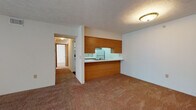 This single-floor, two-bedroom, one-bathroom unit offers a practical and straightforward layout well-suited for everyday living. The living area is carpeted in warm beige tones and features a textured ceiling, with a sliding glass door that opens to a private balcony overlooking lush greenery, bringing in natural light and outdoor views. A half-wall pass-through connects the living space to the kitchen, which is designed in a galley configuration. The kitchen features white cabinetry with wood-tone trim, white appliances including a refrigerator, electric range, and dishwasher, as well as a double sink and dark hardwood-style flooring that adds contrast to the bright cabinetry. Both bedrooms are generously sized, carpeted, and include windows with blinds that allow natural light. One bedroom also features a closet with built-in shelving for added storage. The bathroom is finished with wood-style flooring, a wide vanity with white countertop and a large mirror, and a full bathtub. The entry to the unit includes a closet nearby, providing convenient additional storage. Overall, the space maintains a clean, neutral palette with warm wood accents throughout.