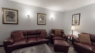 This media room is designed with both comfort and functionality in mind, offering a welcoming space for entertainment and relaxed socializing. The room is furnished with a rich, dark burgundy leather seating suite, including a full-size sofa, loveseat, multiple armchairs with matching ottomans, and accent pillows that add warmth and texture to the ensemble. Plush carpeting underfoot enhances the cozy atmosphere throughout.

A large flat-screen television is prominently wall-mounted and complemented by a dark wood media console below, which features decorative accents and provides convenient storage. The seating is thoughtfully arranged to face the screen, ensuring optimal viewing from every angle.

The light gray walls serve as a neutral backdrop, accented by wall-mounted sconce lighting that creates a warm, ambient glow. Framed artwork depicting tropical palm tree scenes adds a subtle decorative touch. A small bistro-style table with chairs is also present, offering a casual spot for conversation or games.

Black interior doors with clean lines provide a striking contrast to the lighter walls, while ceiling vents and a visible fire safety device reflect practical, well-maintained infrastructure. The overall ambiance is inviting and versatile, suitable for movie nights, social gatherings, or quiet relaxation.