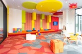 Located on Schering Drive in Union, NJ, this dynamic single-floor community space makes an immediate impression with its bold, contemporary design. The open layout is anchored by a striking geometric patterned carpet in vivid shades of red, coral, orange, and gray — a design element that energizes the entire space and ties its various zones together cohesively.

Floor-to-ceiling windows line multiple walls, inviting abundant natural light throughout and offering open views of the surrounding landscape. A vibrant red accent wall adds a dramatic focal point near the entrance, complemented by a recessed nook with bench seating, perfect for casual gathering or waiting areas. Adjacent glass-partitioned rooms provide semi-private spaces while maintaining visual connectivity to the main area.

A comfortable lounge area features a generous sectional sofa in cool blue-gray tones, paired with a hexagonal coffee table and accented by sculptural floral wall art in warm terracotta tones — adding an artistic, curated touch. A wall-mounted flat-screen display enhances the functionality of this relaxed seating zone.

A dedicated children's area is thoughtfully appointed with small-scale furniture in cheerful orange and white, a whimsical red ceiling light fixture, toys, and colorful play elements. Together, these areas create a welcoming, family-friendly environment designed for community engagement and everyday comfort.