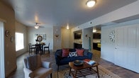 This single-floor residence features an open-concept design that seamlessly connects the living, dining, and kitchen areas into one cohesive and functional space. The living area serves as the central hub, anchored by a navy blue sofa accented with colorful patterned throw pillows and paired with a neutral armchair. A wooden coffee table sits atop a geometric-patterned area rug, adding texture and warmth to the space. Flush-mount ceiling light fixtures provide consistent overhead lighting throughout. The dining area, situated to the left, accommodates a dark wood table and chairs beneath a ceiling fan with a light fixture, complemented by decorative wall art and a round mirror. The kitchen, visible through an open corridor in the background, features dark cabinetry, light countertops, and ample storage space. The entryway is centrally positioned and accessible from the main living area. Throughout the space, light-toned walls contrast with dark wood-finish flooring, creating a balanced aesthetic. The open layout promotes a sense of spaciousness and ease of movement between functional zones.