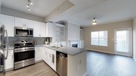 This single-floor residence showcases a thoughtfully designed open-concept layout that seamlessly connects the kitchen and living areas. The kitchen is well-appointed with crisp white cabinetry, granite countertops, and a suite of stainless steel appliances, including a gas range, built-in microwave, and dishwasher. A gray tile backsplash adds a contemporary touch, while the peninsula with an undermount sink serves as both a functional workspace and a natural dividing point between the kitchen and living space. Open shelving built into the dividing wall provides convenient display and storage options. Light wood-tone laminate flooring flows throughout, lending warmth and continuity to the space. The living area benefits from a classic fireplace with a built-in surround, creating a focal point that adds character and comfort. A large window allows generous natural light to fill the room, enhancing the airy, open feel. Neutral wall tones complement the overall palette, contributing to a clean, modern ambiance throughout the home.