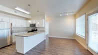 This single-floor space showcases a thoughtfully designed open-concept layout that seamlessly connects the kitchen and living areas. The kitchen is outfitted with crisp white cabinetry, granite countertops, and stainless steel appliances, including a gas range and built-in microwave. A peninsula with an undermount sink serves as both a functional workspace and a natural divider between the kitchen and the adjoining living space. Pendant lighting above the peninsula adds a refined touch, complementing the track lighting installed throughout the main living area. The living room is generously proportioned, featuring warm hardwood-style flooring that runs continuously throughout the space, creating a cohesive and inviting atmosphere. A ceiling fan with an integrated light fixture provides both comfort and additional illumination. Large windows fitted with horizontal blinds allow ample natural light to fill the room, while a glass-paneled door provides access to the exterior. The warm neutral wall tones, textured ceilings, and clean white trim work together to create a bright, welcoming environment that balances contemporary style with everyday functionality.