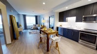 This single-floor residence presents an open-concept design that seamlessly integrates the kitchen, dining, and living areas into one cohesive and functional space. Light wood-toned flooring runs throughout, lending warmth and visual continuity across the entire layout.

The kitchen is outfitted with dark navy cabinetry that extends to the ceiling, providing generous storage while creating a striking contrast against the white countertops and backsplash. A stainless steel sink and modern appliances, including a dishwasher, complete the functional workspace. Recessed lighting illuminates the kitchen area efficiently.

Adjacent to the kitchen, the dining area features a rich wood-toned table paired with contemporary white and natural wood chairs, offering a stylish spot for everyday meals.

The living area occupies the far end of the open plan, anchored by a large window dressed with patterned curtains that allow natural light to brighten the space. A neutral sofa accented with yellow and patterned cushions, a round coffee table, and a layered area rug contribute to a relaxed yet polished atmosphere. A doorway visible in the background suggests access to a separate bedroom, completing this well-appointed layout.
