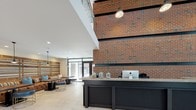 This single-floor space combines contemporary design with distinctive architectural elements to create a polished, functional environment. The lobby serves as the focal point upon entry, anchoring the space with a prominent exposed brick feature wall that rises dramatically, paired with a sleek dark reception desk topped with a stone-look surface. Pendant lighting and recessed fixtures illuminate the area, while a comfortable seating lounge with upholstered furniture and wood-accented walls adds a welcoming atmosphere. Light-toned tile flooring extends throughout the lobby, contributing to a bright, open feel.

Beyond the lobby, a penthouse-level interior room—designated "PH"—offers wood-look plank flooring and floor-to-ceiling black-framed windows and glass doors that flood the space with abundant natural light. This room provides direct access to an expansive outdoor rooftop terrace, fully furnished with modern modular seating arrangements set on large-format light-toned pavers. The terrace is enclosed by horizontal slatted privacy fencing and offers sweeping panoramic views of the surrounding cityscape, making it a standout feature of the overall space.