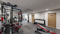 This dedicated fitness facility is thoughtfully designed to support a full spectrum of training needs. The space is divided into distinct functional zones, seamlessly connected to create an efficient workout environment. The strength training area is anchored by a robust multi-station power rack system with cable attachments, barbells, and weight plates, complemented by a well-stocked set of kettlebell racks and dumbbell racks offering a wide range of weights. An adjustable utility bench provides versatility for a variety of exercises. The cardio zone features a treadmill, elliptical trainer, stationary bike, rowing machine, and air bike, catering to all forms of cardiovascular conditioning. A wall-mounted flat-screen television enhances the cardio experience. Mirrored wall panels expand the sense of space and allow for form monitoring. The facility is finished with durable speckled rubber flooring throughout, providing safety and support underfoot. Recessed lighting illuminates the space evenly, while ventilation is ensured through ceiling-mounted HVAC vents. Glass-paneled doors offer visibility between areas, and additional rooms accessible from the main floor provide utility space. Clean white walls and a neutral color palette contribute to a professional, motivating atmosphere.