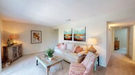 This single-floor residence offers a thoughtfully laid-out living space with a warm, coastal aesthetic carried throughout. The living room serves as a central gathering area, featuring soft carpeted flooring, a textured ceiling, and walls adorned with vibrant teal and earth-toned artwork. A cushioned sofa, a coral-patterned accent chair, and a rustic reclaimed-wood coffee table come together to create a relaxed yet stylish atmosphere.

Adjacent to the living room, the dining area features rich hardwood-style flooring and is anchored by a solid wood table paired with woven rattan chairs. A pendant light overhead and a window with natural light from outside complete the inviting setting.

The kitchen is equipped with dark wood cabinetry, light countertops, and a full range with overhead ventilation, providing functional workspace for everyday cooking.

Two bedrooms offer comfortable retreats, each with plush carpeting, natural light from windows, and warm wood-framed bed frames that echo the coastal theme. One bedroom includes access to a walk-in closet. The two bathrooms are finished with hardwood-style flooring, tub-shower combinations, and vanities with ample counter space, both accented with nautical-themed décor that reinforces the home's cohesive seaside character.