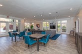 Located at 3800 Club Dr in Norcross, Georgia, this single-floor community clubhouse offers a well-appointed gathering space designed for both relaxation and social interaction. The open-plan layout flows seamlessly between a generously sized lounge area and a dedicated dining zone, all finished with light wood-tone flooring and crisp crown molding throughout.

The lounge area centers around a large sectional sofa accented with colorful throw pillows, complemented by accent chairs and a coffee table set atop a rich blue area rug. A built-in media wall with shelving and a flat-screen television anchors one end of the room, while an adjacent pass-through opening connects to a full kitchen with bar seating. Large floor-to-ceiling windows and glass doors flood the space with natural light and provide direct access to an outdoor patio, offering pleasant views of the surrounding greenery.

The dining area features a bold navy blue accent wall adorned with vibrant abstract artwork, creating a striking visual contrast against the lighter tones of the rest of the space. A dining table with teal upholstered chairs provides a functional and stylish setting. French doors with glass panels lead to a stairwell, adding architectural character. Recessed lighting throughout ensures a bright, welcoming ambiance, making this clubhouse an inviting hub for community gatherings.