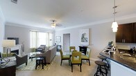 This single-floor residence is thoughtfully designed with an open, flowing layout that seamlessly connects the main living areas. The entryway, finished with hardwood-style flooring and crisp white crown molding, provides a welcoming introduction to the space, with clear sightlines into the adjoining rooms.

The combined living and dining area is generously proportioned, featuring plush wall-to-wall carpeting, smooth ceilings with crown molding detail, and a ceiling fan with integrated lighting. Large windows dressed with green sheer curtains allow natural light to fill the space, creating a bright and airy atmosphere. The dining area accommodates a glass-top table surrounded by green upholstered chairs, while the living area offers comfortable seating arranged around a patterned area rug.

To one side, the kitchen features dark espresso cabinetry with ample storage, dark countertops, and a sink with adjacent counter seating, offering both functionality and style. A secondary room off the hallway, visible through an open doorway, appears to serve as an additional bedroom or den. Throughout, the warm beige and white color palette, consistent trim work, and cohesive finishes contribute to a polished, unified aesthetic.