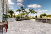 Located at Presidential Way in West Palm Beach, Florida, this single-level outdoor amenity space is thoughtfully designed to deliver a resort-style experience within a well-appointed residential community. The grounds are finished with large-format stone pavers throughout, providing a cohesive and polished aesthetic that anchors the entire outdoor environment.

At the heart of the space is a generously sized swimming pool with crystal-clear water, framed by tropical palm trees, manicured plantings, and lounge seating — all enclosed by a classic white metal fence that adds structure while preserving open sightlines. Adjacent to the pool, a full outdoor bar with bar-height seating offers a dedicated social gathering area, complemented by Adirondack chairs set along a lush green lawn.

The surrounding multi-story residential building features a classic coastal architectural style, with arched entryways, stone-clad base detailing, white-framed windows, and private balconies. Tropical landscaping — including tall royal palms, flowering shrubs, and layered greenery — lines the perimeter, creating a lush, garden-like atmosphere. The open-air design takes full advantage of the South Florida climate, offering abundant natural light and sweeping views of the surrounding skyline and landscape beneath wide, sun-filled skies.