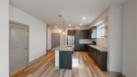 This single-floor residence showcases a contemporary design with a cohesive mix of materials and finishes throughout. The kitchen serves as a central highlight, featuring dark espresso-toned cabinetry that extends to the ceiling, granite countertops, and a white subway tile backsplash. A central island with a built-in sink provides additional prep space and seating potential, while pendant lighting above adds a refined touch. Stainless steel appliances, including a full-size refrigerator and range, complete the functional layout. Wood-look plank flooring runs throughout the main living areas, lending warmth and continuity to the space.

The living area is bright and open, benefiting from multiple windows fitted with blinds that allow natural light to filter in. Neutral wall tones complement the flooring and trim details throughout. A bedroom area offers plush carpeting and ample natural light through well-positioned windows.

Storage is well-addressed with a generously sized walk-in closet outfitted with multi-tier wire shelving on three walls, providing extensive organizational options. The bathroom features large-format light tile flooring, a dark vanity with granite countertop, an undermount sink, and a well-lit mirror. The overall aesthetic is clean, modern, and move-in ready.