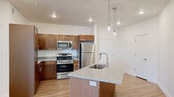 This well-appointed single-floor residence offers a thoughtfully designed open-concept layout that seamlessly connects the kitchen and living areas. The kitchen is a standout feature, showcasing rich warm-toned wood cabinetry, granite countertops, and a full suite of stainless steel appliances including a gas range, microwave, and refrigerator. A central island with an integrated sink and additional granite surface space serves as both a functional workspace and a natural gathering point, illuminated by stylish pendant lighting suspended from above. Light wood-style flooring runs throughout the main living areas, contributing to a clean and contemporary aesthetic.

The bedrooms offer a contrast in comfort, featuring plush carpeted flooring, neutral wall tones, and window blinds that allow for natural light. One living area opens directly onto a private balcony through sliding glass doors, providing elevated panoramic views of the surrounding cityscape and neighborhood rooftops — a notable highlight of the space.

The bathroom continues the refined finishes seen throughout, with a granite-topped vanity, an undermount sink, and a large mirror. Clean white walls and recessed lighting maintain the unit's bright and cohesive design language. Overall, this residence combines functional, modern finishes with a comfortable and airy atmosphere.