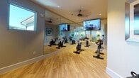 This fitness center is thoughtfully designed to accommodate a variety of workout styles within a single-level layout. The space is divided into distinct functional zones, each outfitted with purpose-specific equipment. The cycling area serves as a centerpiece, featuring multiple stationary bikes arranged before a full wall of floor-to-ceiling mirrors that amplify the sense of space. Mounted flat-screen televisions and a ceiling fan add comfort and entertainment during workouts. Recessed lighting and natural light from horizontal windows along the upper walls keep the environment bright and energetic.

A dedicated functional training zone houses a well-organized rack of kettlebells in graduated sizes, displayed beneath two framed contemporary art pieces with silver metallic frames, lending a polished aesthetic to the space. Adjacent areas provide room for stretching and group fitness activities, complete with stability balls, medicine balls, yoga mats, and foam rollers neatly stored on a Smart Fitness rack.

Entry to the space is through sleek double doors featuring full-length glass panels, contributing to the open and modern feel. Light wood-look flooring runs throughout, tying the zones together cohesively. The overall ambiance is clean, modern, and welcoming.