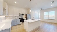 This thoughtfully designed 1-bedroom, 1-bathroom unit offers a bright and contemporary living experience across a single floor. Light wood flooring flows seamlessly throughout, creating a cohesive, airy aesthetic. The open-concept kitchen is a standout feature, showcasing a large center island with an integrated sink and pendant lighting above, two-tone cabinetry with upper white shaker-style cabinets and lower gray cabinetry, a white quartz countertop, a marble-patterned hexagonal tile backsplash, and a full suite of stainless steel appliances including a range, microwave, and refrigerator. The kitchen opens directly into the living area, which is illuminated by natural light from a large window fitted with blinds and equipped with a ceiling fan. The bedroom is a well-proportioned, comfortable retreat with its own window for natural light, a ceiling fan, and direct access to a dedicated closet. The bathroom features a soaking tub surrounded by floor-to-ceiling marble-look tile with an elegant mosaic accent border. A dedicated in-unit laundry area houses a full-size side-by-side washer and dryer with overhead shelving, adding everyday convenience to this move-in ready space.
