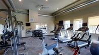 This fitness center offers a comprehensive workout environment designed to accommodate a variety of exercise routines. The space is outfitted with multiple cardio machines, including treadmills and stationary bikes, all positioned to take advantage of natural light streaming in through large windows along the exterior wall. A full-length mirrored wall expands the sense of space and allows exercisers to monitor their form during workouts. A dedicated free weights area features a well-stocked dumbbell rack, providing options for strength training alongside the cardio equipment. Each cardio machine is equipped with an individual monitor for entertainment during workouts. The room is finished with durable, patterned carpet flooring suited to high-traffic athletic use, while exposed ductwork and a ceiling fan contribute to an industrial-modern aesthetic. Recessed lighting and the ceiling fan work together to maintain a comfortable, well-lit environment throughout. The neutral wall tones and functional layout create a clean, focused atmosphere conducive to productive training sessions. Overall, the fitness center is thoughtfully designed to meet the needs of a wide range of fitness enthusiasts.