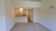This two-story space is defined by its dramatic double-height ceilings in the main living area, creating an airy, open atmosphere. A standout feature is the breakfast bar, which is adorned with a decorative stacked-stone facade and topped with a granite-style surface, serving as a natural divider between the carpeted living area and the kitchen beyond. The kitchen is equipped with white cabinetry, a microwave, and a full-size refrigerator, with a ceiling fan providing overhead lighting and airflow. Adjacent to the kitchen, a hallway leads to additional rooms, flanked by tall white cabinetry offering generous storage. The living area features neutral wall-to-wall carpeting that contributes to a warm, understated aesthetic. Above, a loft level is accessible via a staircase and is visible from the main floor through an open railing, adding functional square footage and architectural interest. The predominantly white color palette throughout enhances the sense of brightness and space, making the interior feel clean and open.