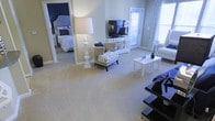 This single-floor space features an open and airy living area centered around plush, neutral-toned wall-to-wall carpeting that creates a warm, cohesive foundation throughout. The living area is generously sized, accommodating a comfortable seating arrangement that includes a classic chaise lounge with a bold zebra-print pattern and a sofa, complemented by a sleek white coffee table. A tall, sculptural floor lamp adds an elegant focal point to the room.

Natural light flows in through multiple windows dressed with patterned curtains, lending a bright and welcoming atmosphere. A media console positioned against the wall houses a flat-screen television, while a door nearby provides access to an outdoor area.

Directly accessible from the living space is a bedroom featuring a deep navy accent wall, a tufted upholstered headboard, and coordinating blue and white bedding, creating a stylish, cohesive color palette that connects the two spaces. The navy and white tones used throughout the bedroom are echoed in the living area's accent pieces, giving the overall space a well-designed, harmonious aesthetic.