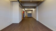 Located at 230 Perry St in Buffalo, NY, this single-floor unit blends industrial character with modern updates throughout its layout. The main living area is expansive and elongated, featuring rich dark hardwood-style flooring, exposed ceiling ductwork, and structural columns that contribute to its loft-like aesthetic. Track and ceiling-mounted lighting illuminate the space efficiently, complementing the neutral gray and white walls.

A standout feature is the kitchen, which makes a bold visual statement with high-gloss red cabinetry, dark countertops, and stainless steel appliances including a gas range, dishwasher, and refrigerator. An open ceiling with black-painted infrastructure above the kitchen adds to the edgy, urban feel.

Toward the front of the unit, a separate room features double glass entry doors and a large window, allowing natural light to filter in and offering a flexible space suitable for an office, studio, or additional living area. A bathroom with light tile flooring, clean white walls, and practical fixtures provides functional utility.

The floor plan model reveals multiple distinct zones, allowing for versatile use of the space. Overall, the unit delivers an urban industrial ambiance with thoughtful modern finishes, ideal for those seeking a dynamic, loft-inspired environment in Buffalo.