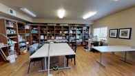 This dedicated craft room is a generously sized, single-floor space designed to accommodate a variety of creative pursuits. The room features warm-toned walls and light hardwood flooring throughout, creating a bright and welcoming environment conducive to focused work.

Central to the layout are multiple large, adjustable work tables that provide ample surface area for cutting, sewing, and assembling projects. The tables are arranged to allow comfortable movement around each workstation. Along the back wall, an extensive run of open wooden shelving units spans nearly the full width of the room, offering substantial organized storage for craft supplies, bins, and materials.

Natural light enters through several windows fitted with horizontal blinds, supplemented by multiple overhead fluorescent light fixtures that ensure consistent, well-distributed illumination throughout the space. Framed artwork adorns the walls, adding a personal and creative touch to the room's ambiance.

A dedicated sewing area is visible along one wall, equipped with sewing machines and additional worktables. A utility area along one side of the room includes medium-toned wood cabinetry, upper and lower storage, a sink, and counter space, providing practical support for hands-on crafting needs. A private door leads to an adjacent space. The overall layout is functional, organized, and well-suited for both individual and group crafting activities.