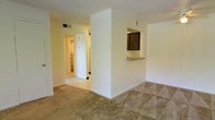 This single-floor residential space offers a functional and straightforward layout designed for everyday comfort. The living area is finished with wall-to-wall carpeting in a neutral tone, providing a warm and understated foundation throughout the main space. A defining architectural feature is the partial dividing wall with a pass-through window opening into the kitchen, accented by a wooden cabinet above — a classic detail that connects the two spaces while maintaining a sense of separation. The kitchen pass-through also includes a small countertop ledge, ideal for casual dining or convenient serving. The walls are finished in a soft, light tone, contributing to a bright and airy atmosphere, while the textured popcorn ceiling adds a layer of visual detail consistent with the building's era of construction. Overhead HVAC vents are integrated into both the ceiling and upper walls, ensuring climate control throughout. A hallway visible from the main living space leads to additional rooms, hinting at a practical and connected floor plan. The overall ambiance is clean, neutral, and move-in ready, offering a blank canvas well-suited for personalization.