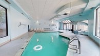 This indoor pool facility offers a welcoming aquatic environment designed for both leisure and relaxation. The centerpiece is a generously sized swimming pool with clear, turquoise water, equipped with stainless steel entry rails for easy access. Adjacent to the main pool is a round hot tub, providing a dedicated space for hydrotherapy and unwinding.

The surrounding deck area is well-appointed with rows of white lounge chairs, allowing guests to comfortably rest poolside. A seating area with tables and chairs is positioned near a bright, windowed glass enclosure at the far end of the space, inviting natural light into the room and offering a pleasant atmosphere for relaxation.

The interior is finished with light blue walls and a drop-panel ceiling fitted with recessed lighting, creating a clean and airy feel throughout. Decorative patio umbrellas add a resort-like charm to the space. A wall clock and informational signage are also present, ensuring guests have all the practical amenities they need. The overall ambiance is calm and inviting, making this indoor pool area a comfortable destination for aquatic recreation year-round.