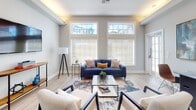 Located at 3415 SW 39th Blvd in Gainesville, FL, this single-floor space offers a thoughtfully designed interior with a cohesive modern aesthetic. The living area is a standout feature, boasting elevated ceilings with warm tray lighting that adds a welcoming glow. A pair of large, grid-paned windows flood the room with natural light while offering views of the surrounding neighborhood. Light wood-tone flooring runs throughout, complementing the neutral gray walls and crisp white trim. The living area is anchored by a bold blue sofa, a glass-top coffee table, and a mounted flat-screen television, creating a comfortable and functional gathering space. Adjacent to the living area is a dedicated office space that continues the blue-and-gray color palette. This room features a long, sleek workstation with multiple monitors, a seating area with plush blue accent chairs, abstract wall art, and a tray ceiling with recessed lighting — making it ideal for productive work. A cozy reading nook with a blue velvet chair, a round wall mirror, and curated decor adds an inviting touch between spaces. Throughout the interior, light hardwood floors, bold blue accents, and carefully chosen artwork create a polished, modern atmosphere that is both functional and visually engaging.