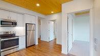 This single-floor unit showcases a thoughtfully designed interior where natural wood plank ceilings serve as a defining architectural feature throughout the main living and kitchen areas, adding warmth against crisp white walls and recessed lighting. The kitchen is well-appointed with flat-front white cabinetry extending to the ceiling, light-toned countertops, a white subway tile backsplash with under-cabinet lighting, and a full suite of stainless steel appliances including a refrigerator, electric range, over-the-range microwave, and dishwasher. Wood-look flooring flows consistently through the main areas, reinforcing a cohesive, contemporary aesthetic. The living area benefits from generous natural light through multiple large windows fitted with horizontal blinds, offering treetop views and a ceiling fan overhead. A dedicated laundry area houses a stacked washer and dryer unit. The bathroom features a tub-shower combination, wood-look flooring, built-in shelving, and clean white finishes. Storage is well-addressed with a spacious walk-in closet equipped with wire shelving systems and carpeted flooring. Overall, the unit presents a clean, modern atmosphere with warm natural accents throughout.